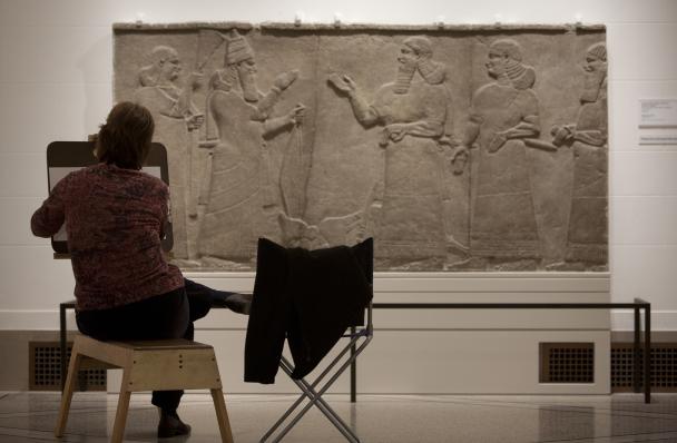 A visitor sits beside a stool, drawing on a bench in the Ancient Middle East galleries at the DIA